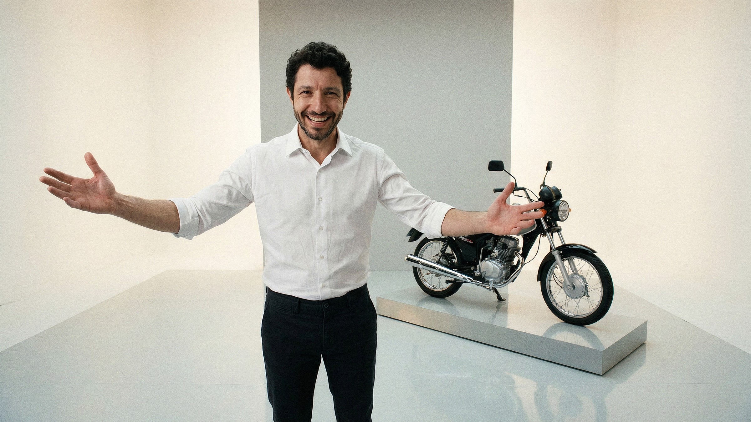 Comercial de varejo: ele de braços abertos e sorriso enorme para a lente, a moto exposta num praticável ao lado, ciclorama claro.