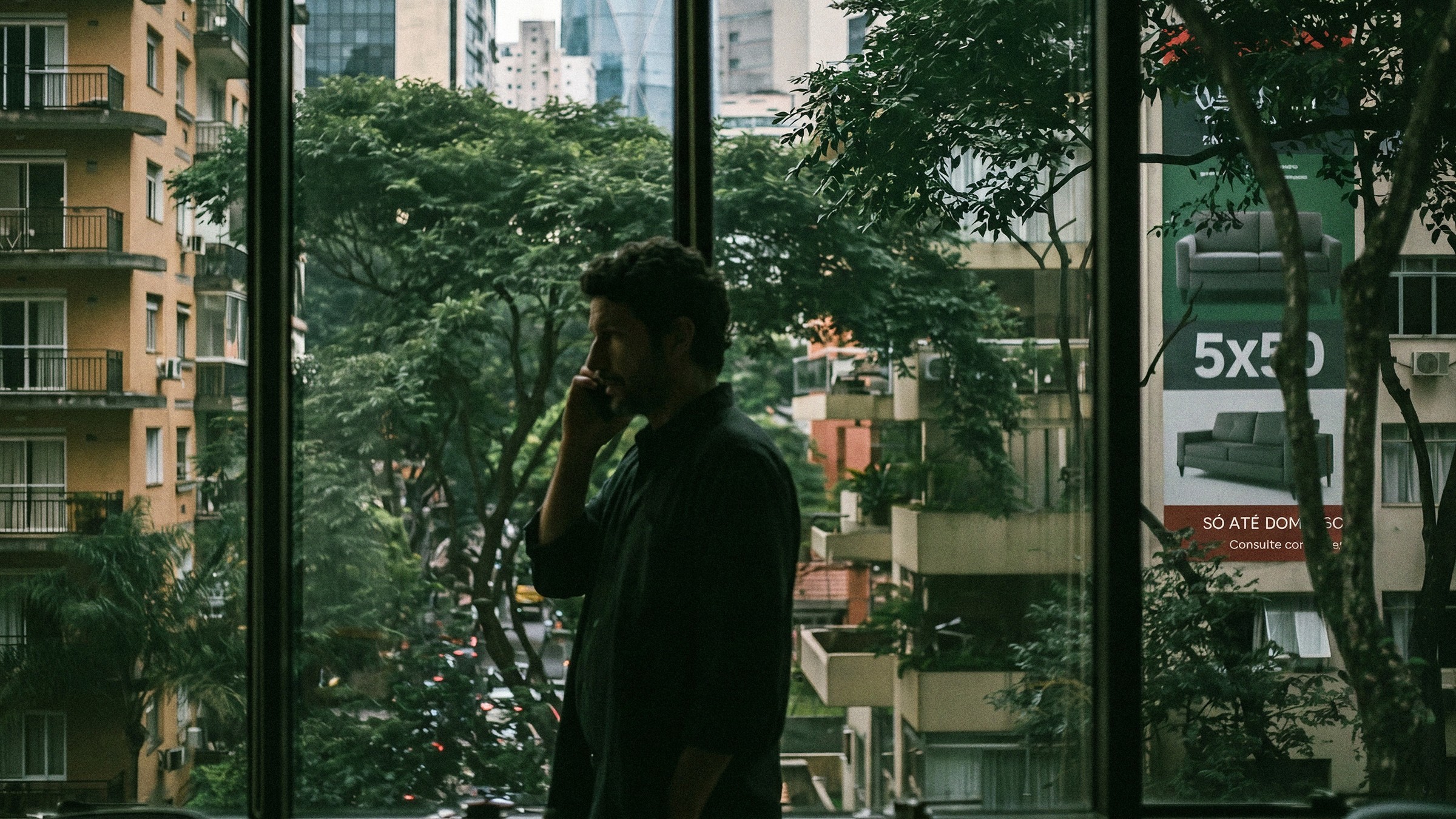 Ele ao telefone contra a janela, e do lado de fora um outdoor de varejo com 5x de R$ 50.