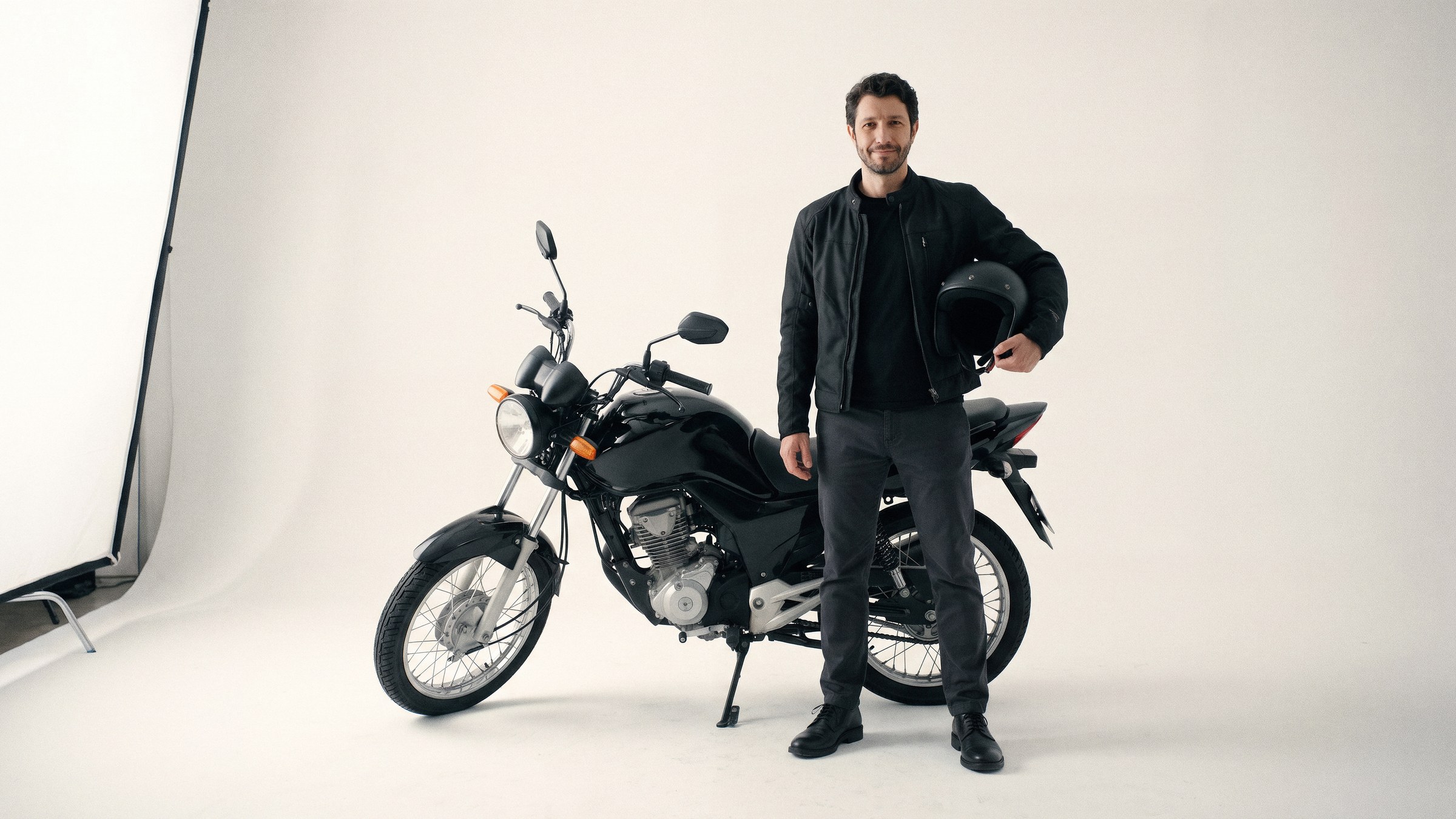 Ele em pé ao lado de uma moto, jaqueta e capacete no braço, sem gesto de apresentação.