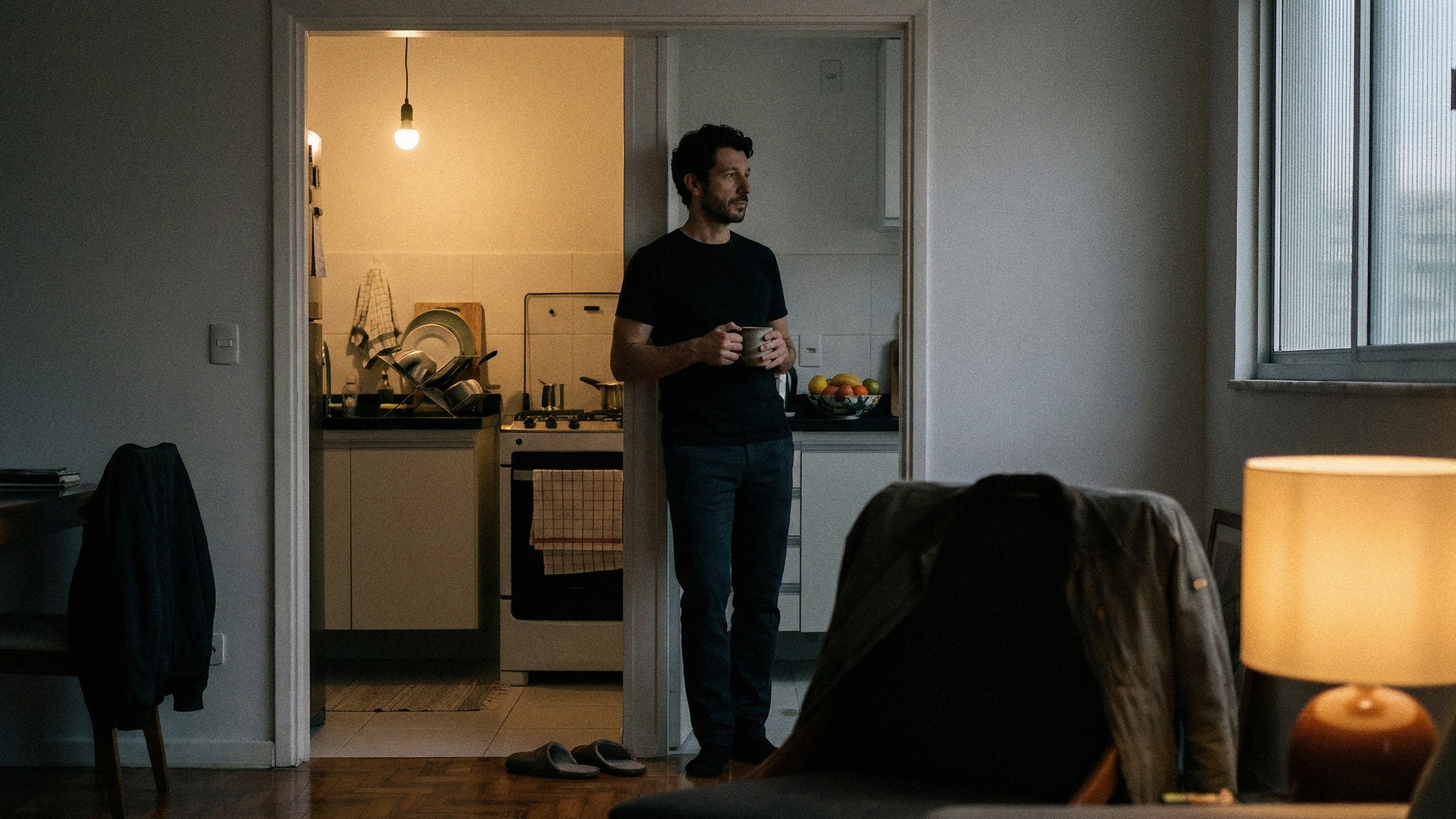 A casa dele à noite: ele parado na porta da cozinha com uma caneca, louça secando, jaqueta na cadeira, abajur aceso na sala.